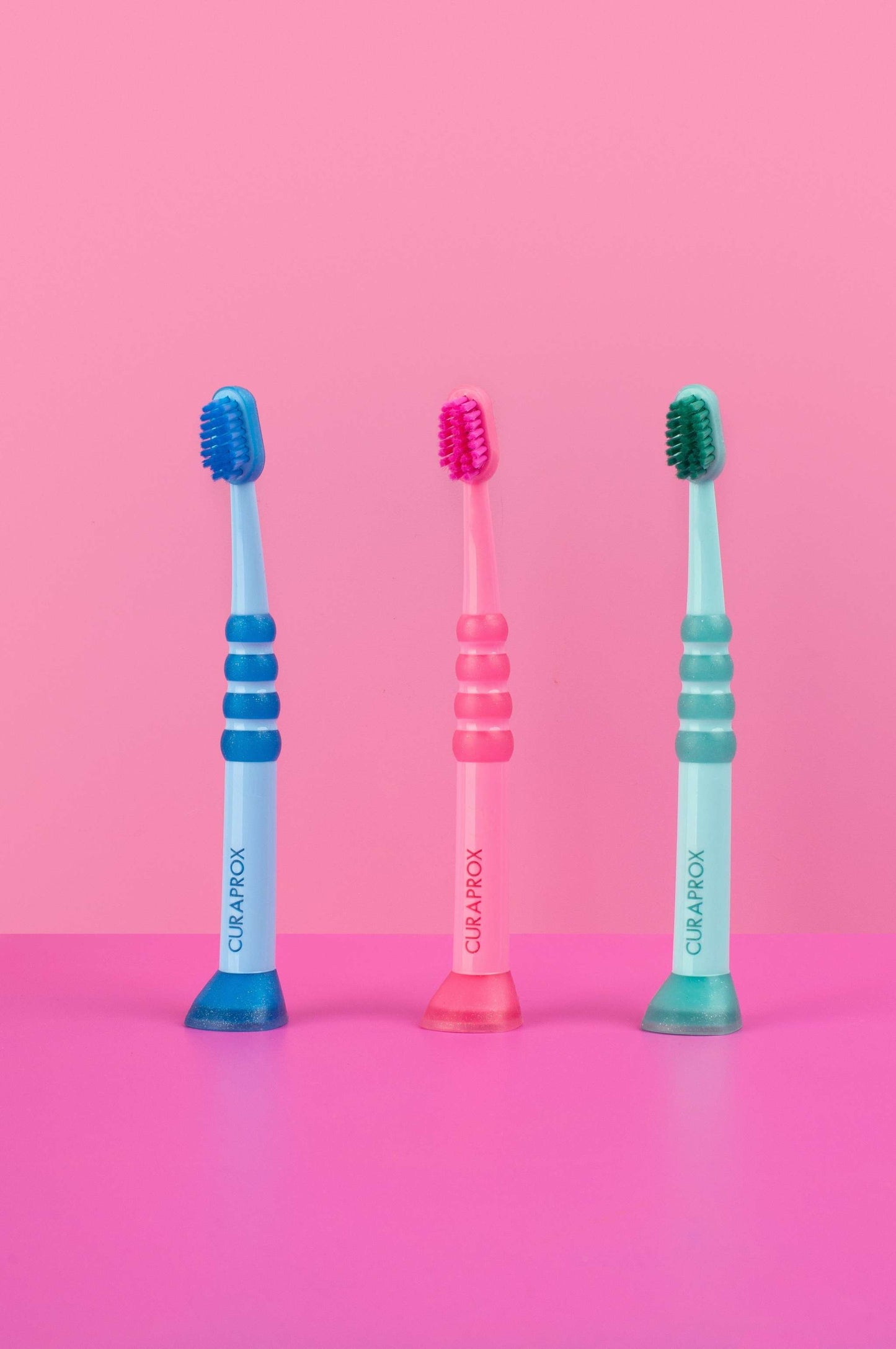 Three Curaprox baby toothbrushes for ages 0-4 years in blue, pink, and green with rubberized handles against a pink background