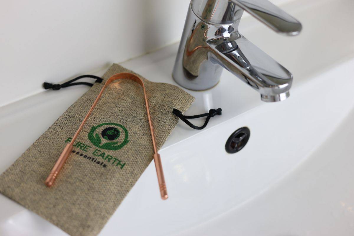 Copper tongue scraper with thick handles and wide scraping edge placed on a linen carry bag beside a bathroom sink faucet