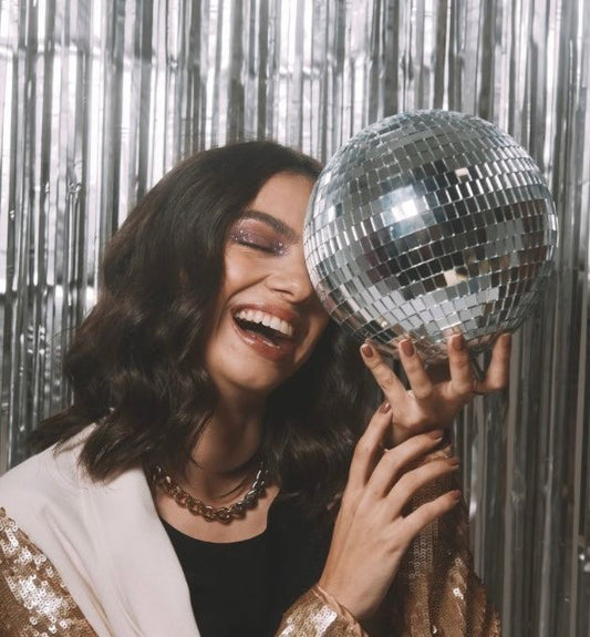 Smiling woman holding disco ball celebrating New Year with joyful expression and shiny backdrop, representing 2026 oral health resolutions.