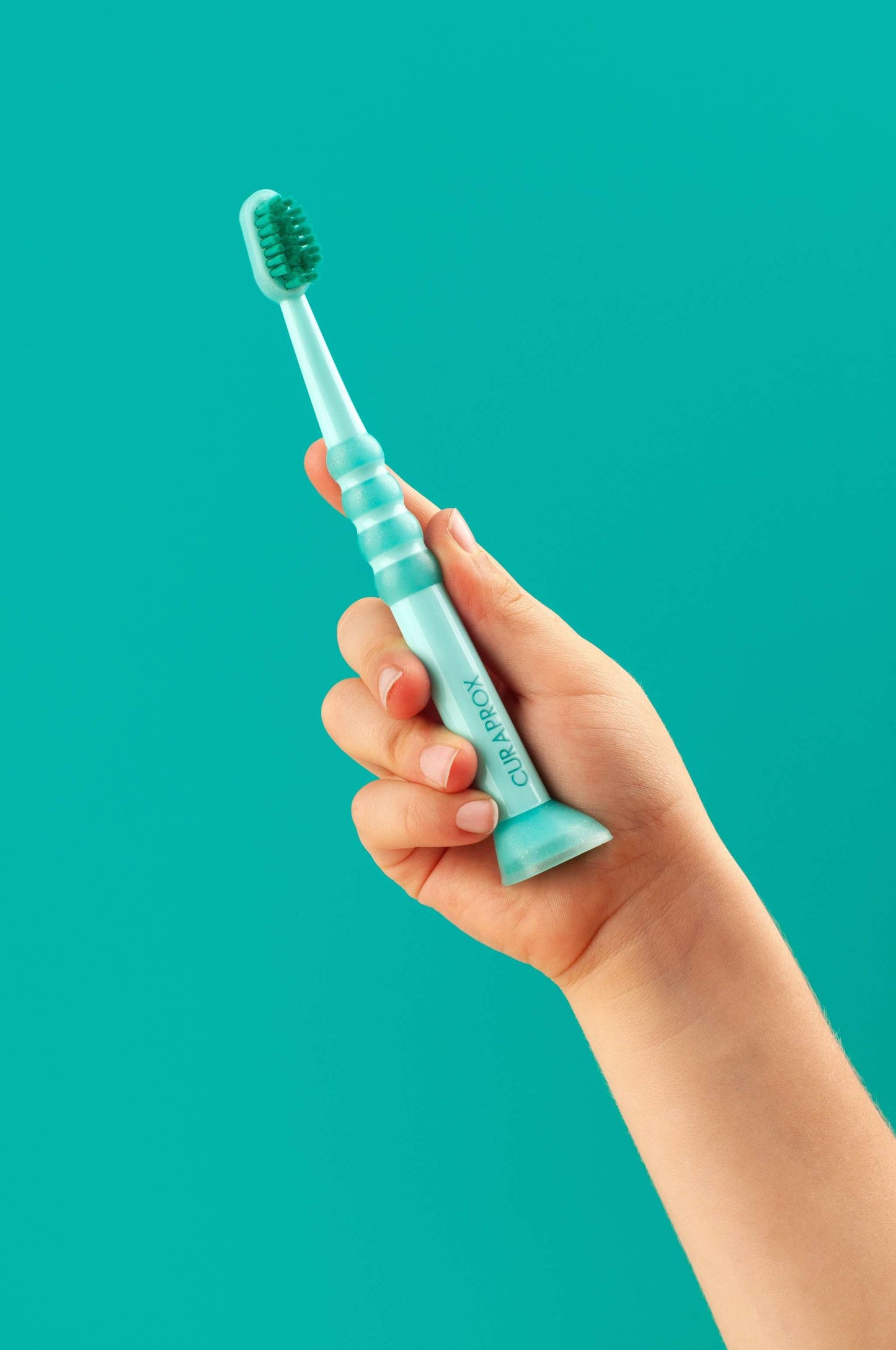 Curaprox Baby Toothbrush for 0-4 years with fine filaments, rubberized small brush head, and anti-slip handle held by a child's hand on teal background