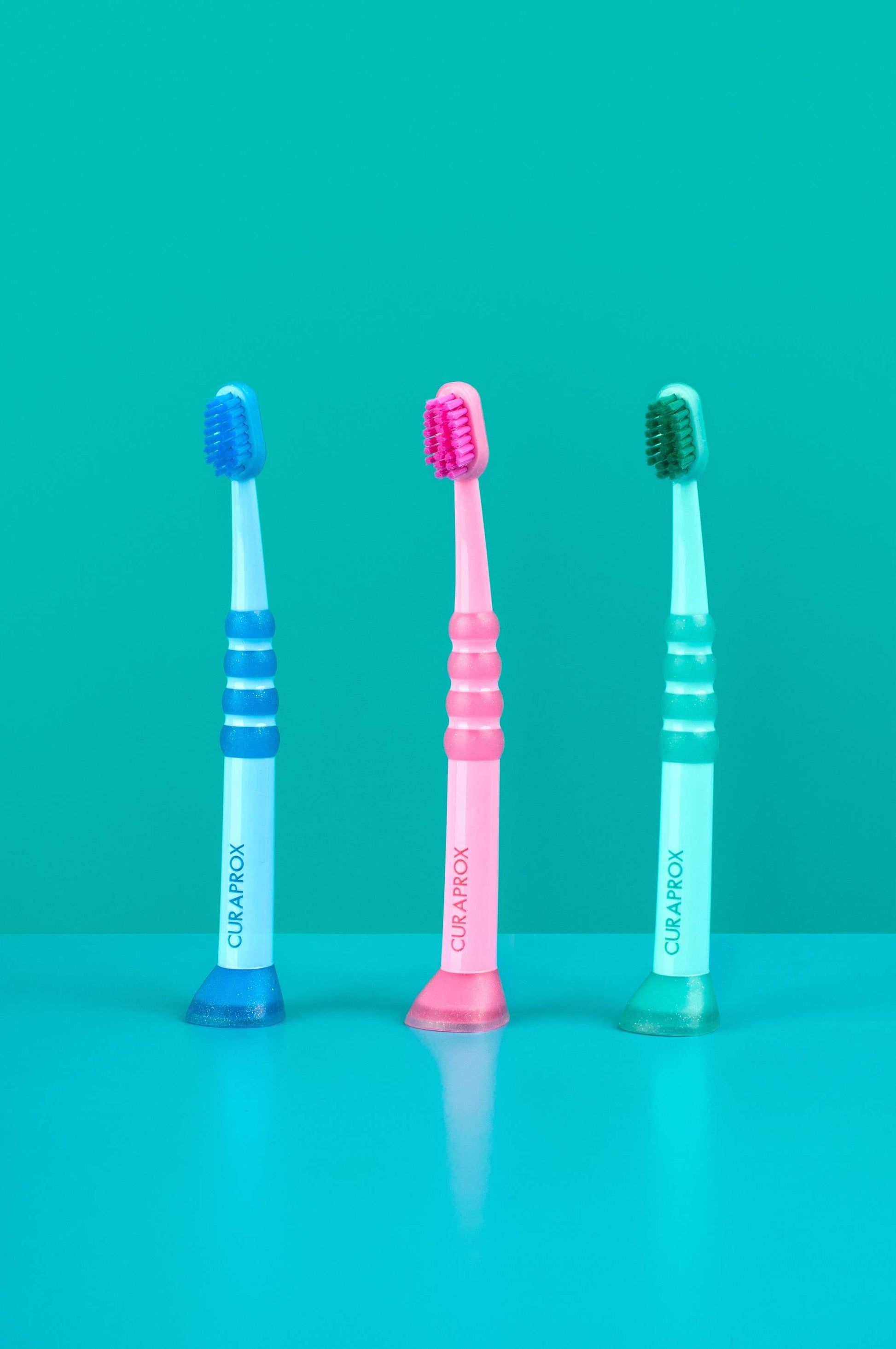 Three Curaprox Baby Toothbrushes for ages 0-4 in blue, pink, and green with fine filaments and anti-slip handles.