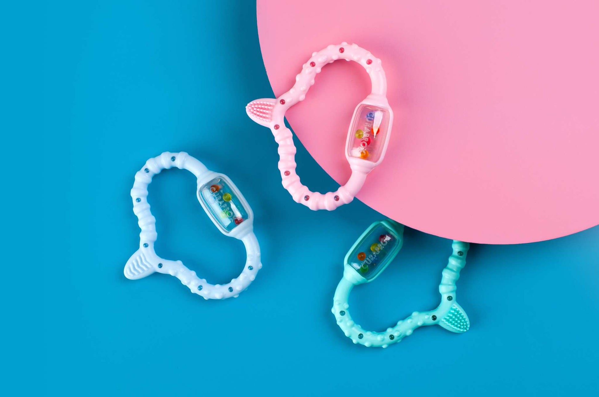 Curaprox Baby teething rings in pink, blue, and teal colors on blue and pink background