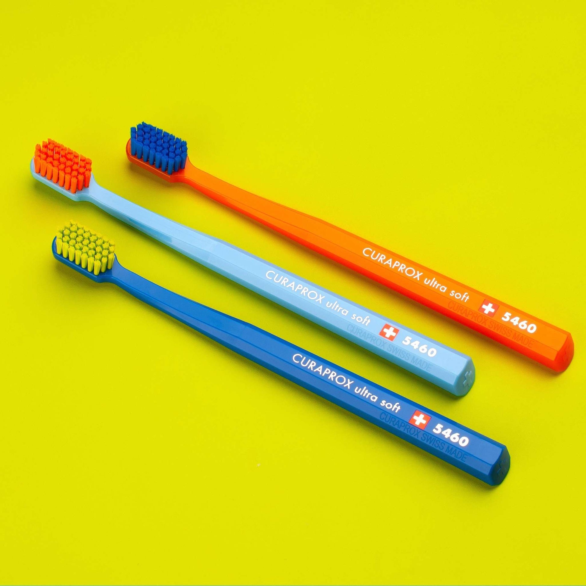 Set of three Curaprox CS 5460 ultra soft toothbrushes in bright blue, light blue, and orange colors on yellow background