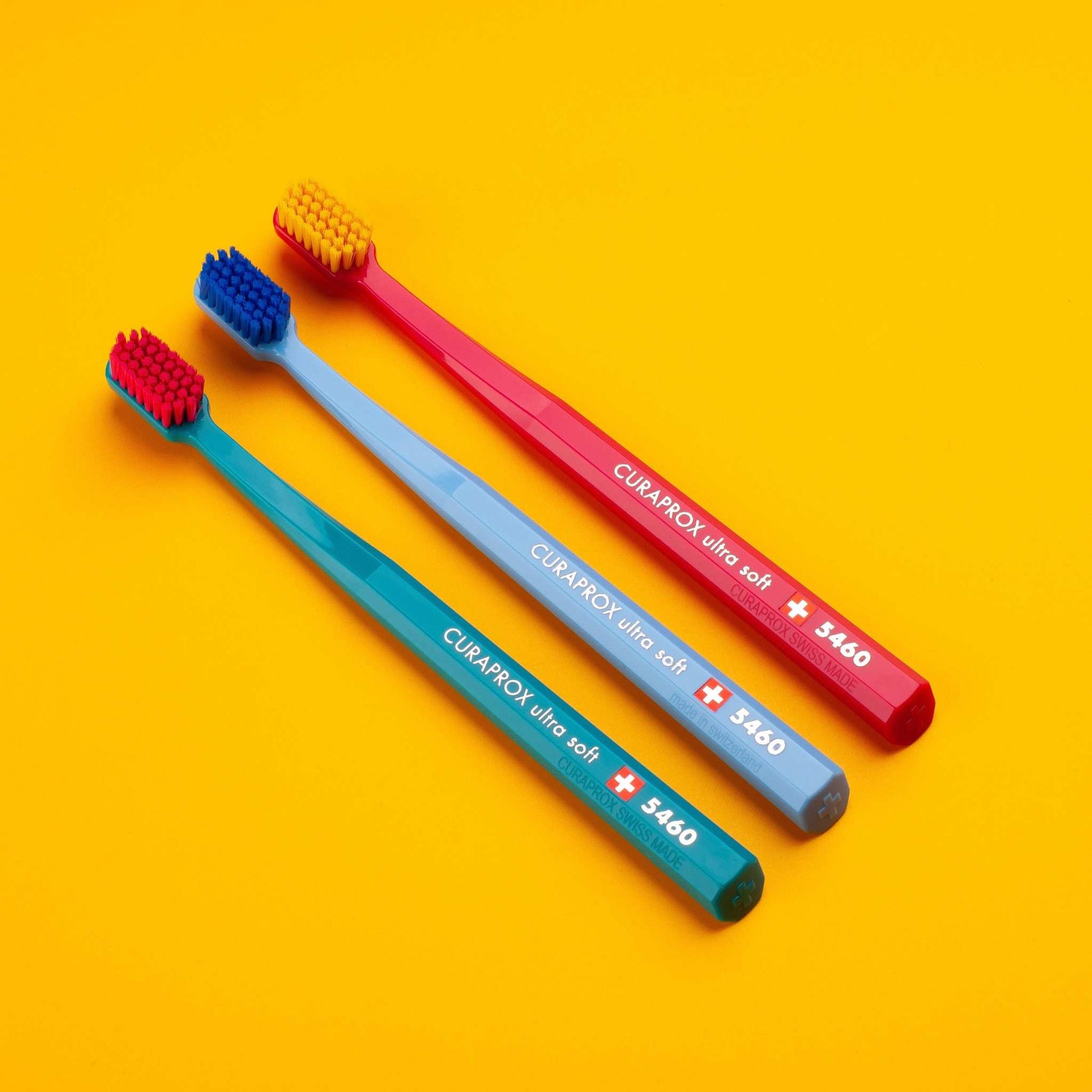 Three Curaprox CS 5460 ultra soft toothbrushes in red, blue, and teal on a yellow background