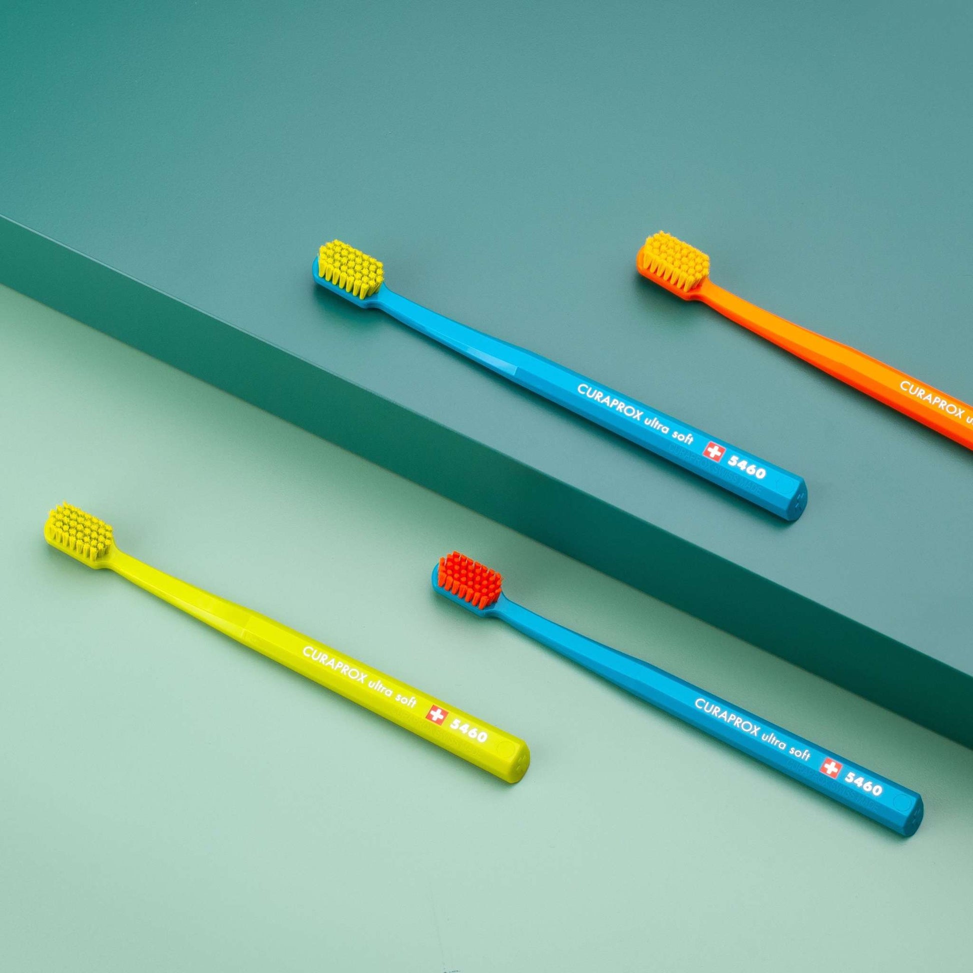 Four Curaprox CS 5460 ultra soft toothbrushes in assorted colors with angled heads and dense bristles on a two-tone green background.