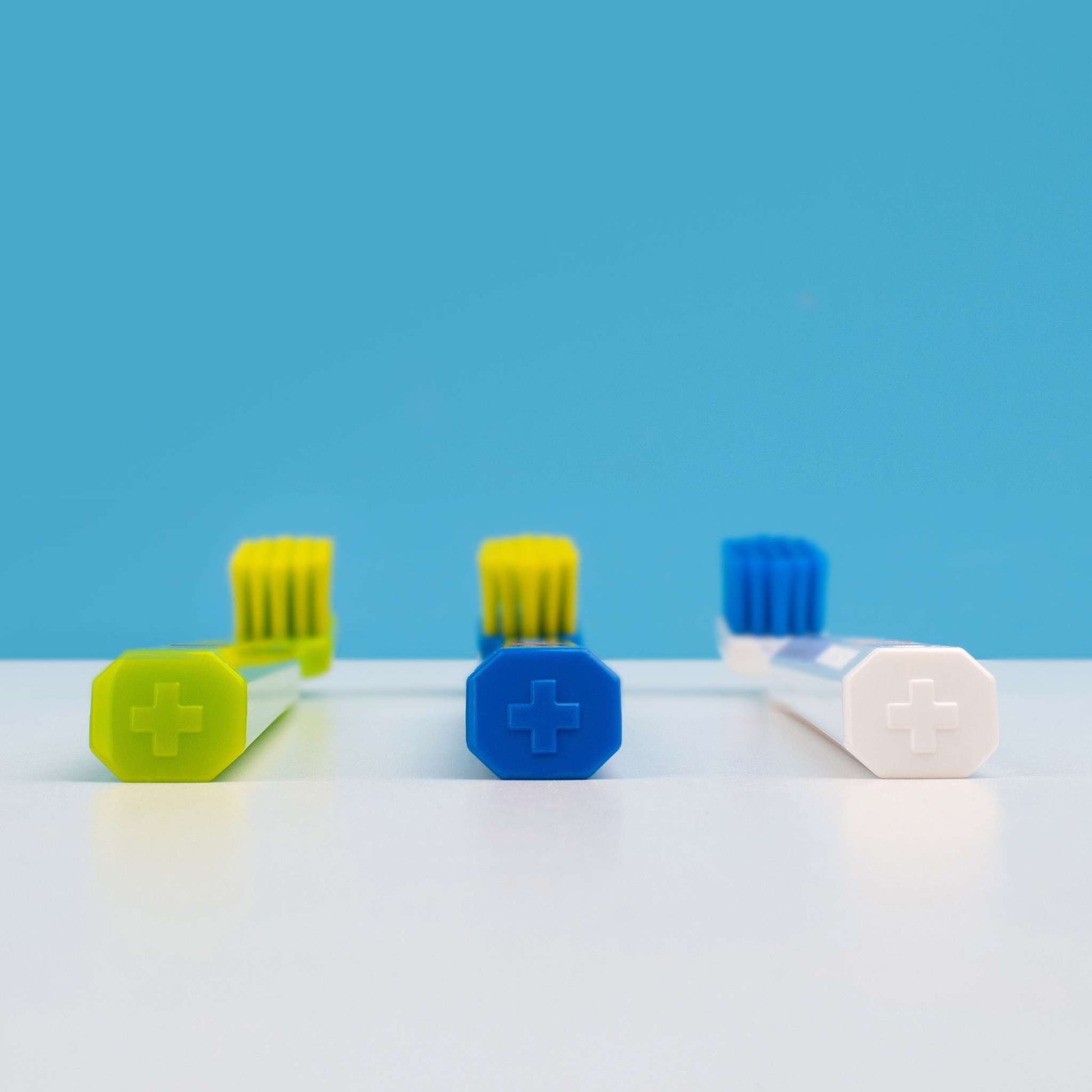 Three Curaprox CS 5460 ultra soft toothbrushes with octagonal handles in green, blue, and white colors against a blue background.
