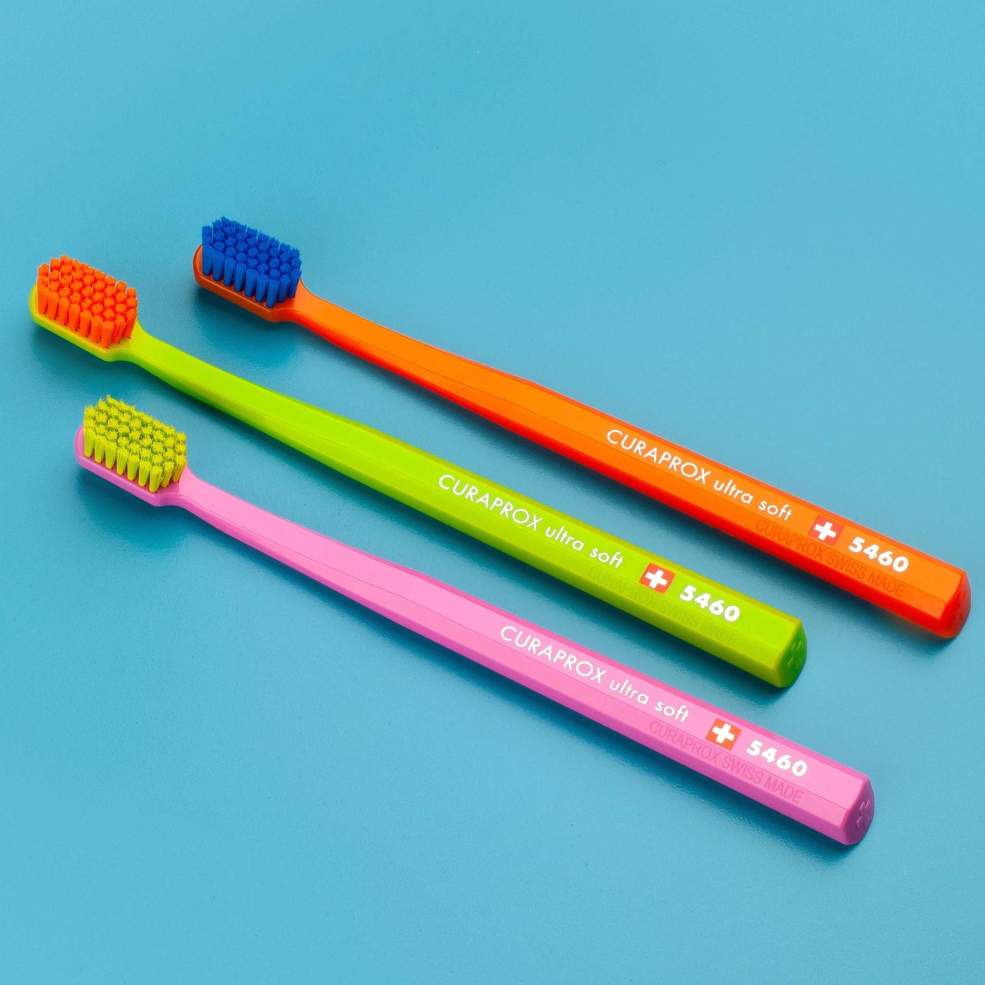 Three Curaprox CS 5460 ultra soft toothbrushes in orange, green, and pink handles with colorful bristles on a blue background
