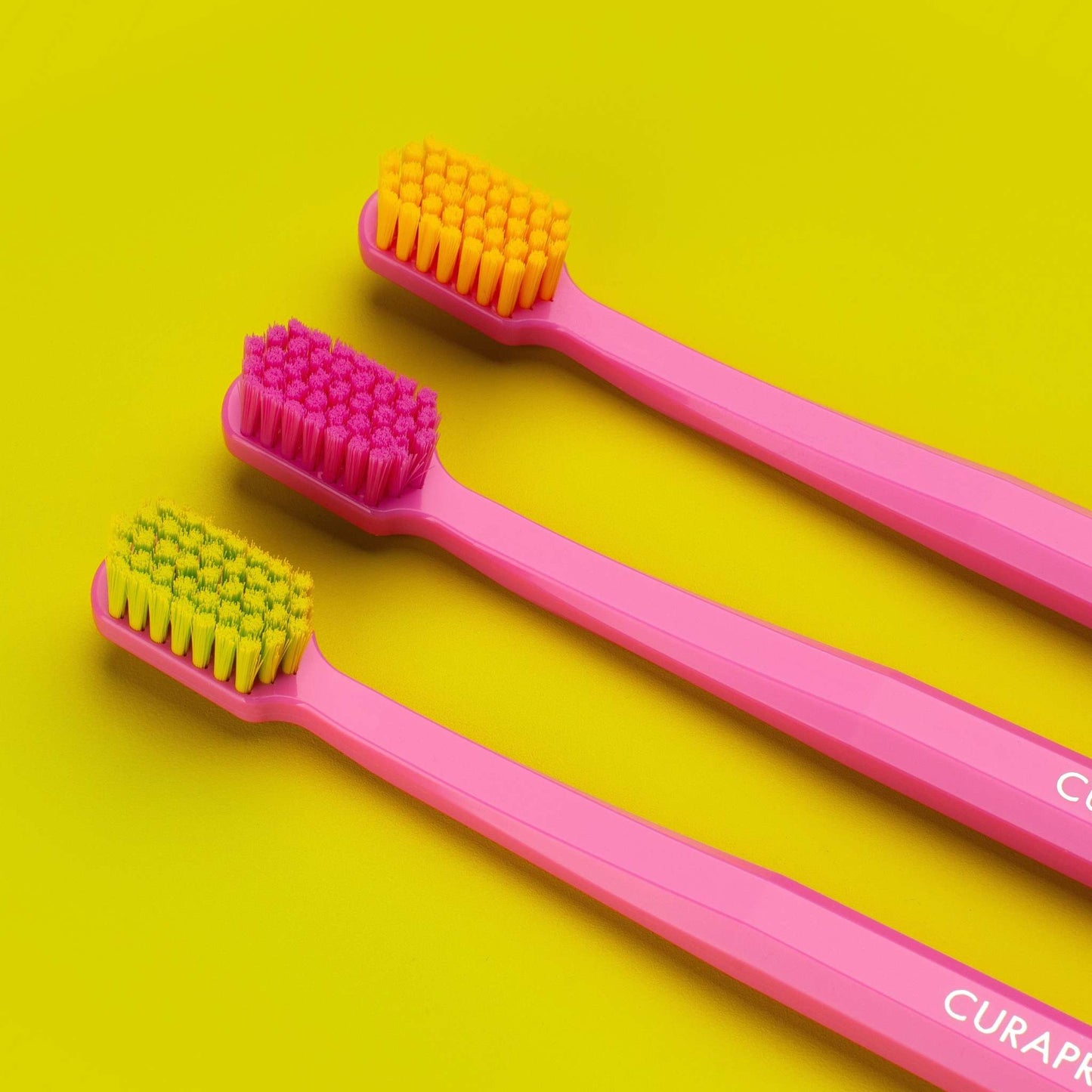 Set of three Curaprox CS 5460 ultra soft toothbrushes with densely packed colorful bristles on pink octagonal handles against a yellow background