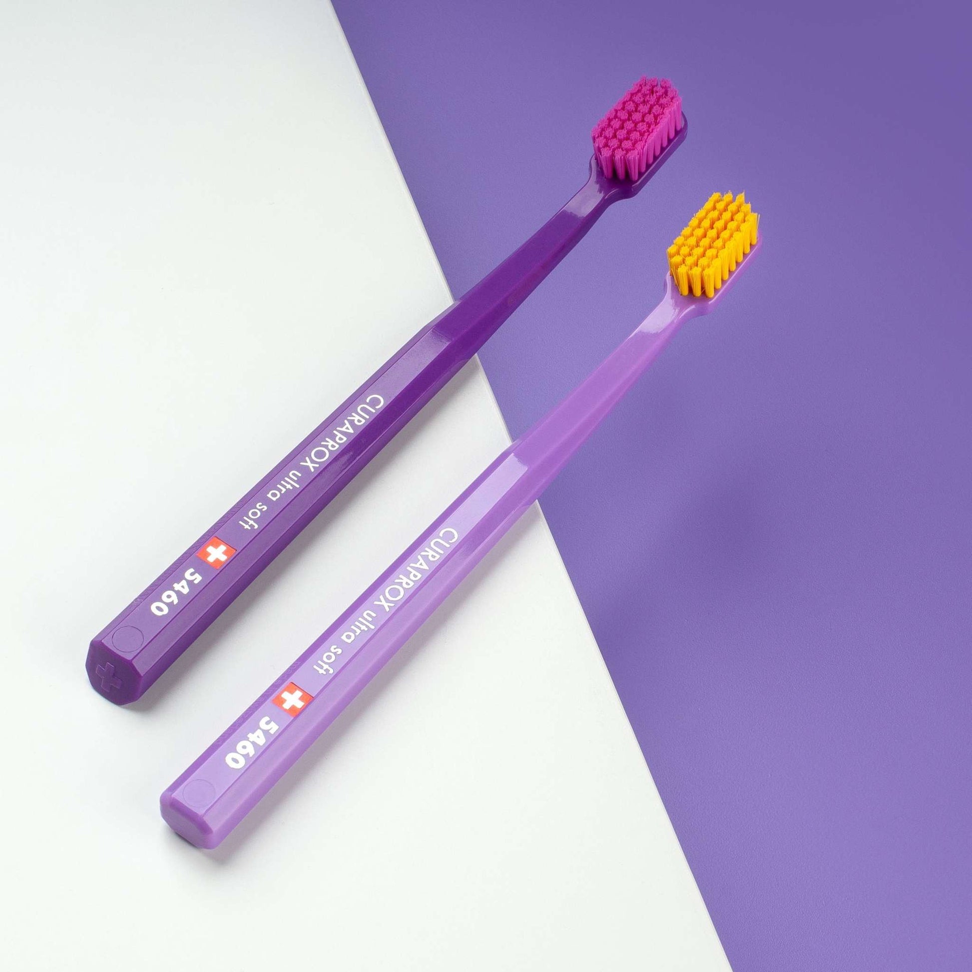 Two Curaprox CS 5460 ultra soft toothbrushes with dense bristles, one with a purple handle and pink head, the other lavender with an orange head, displayed on a white and purple background.